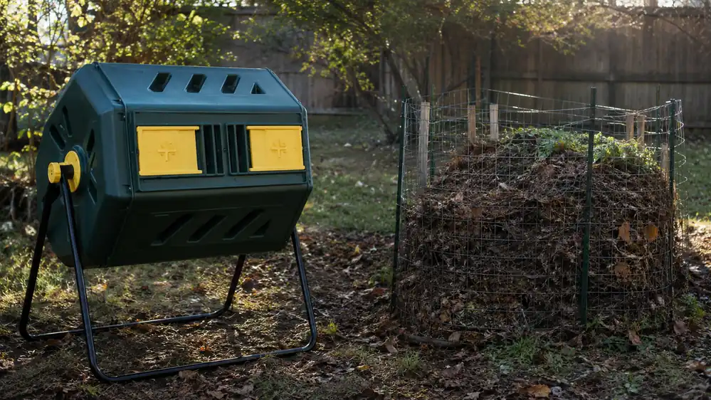 A black dual-chamber compost tumbler on a steel frame standing next to an open wire compost pile in a backyard
