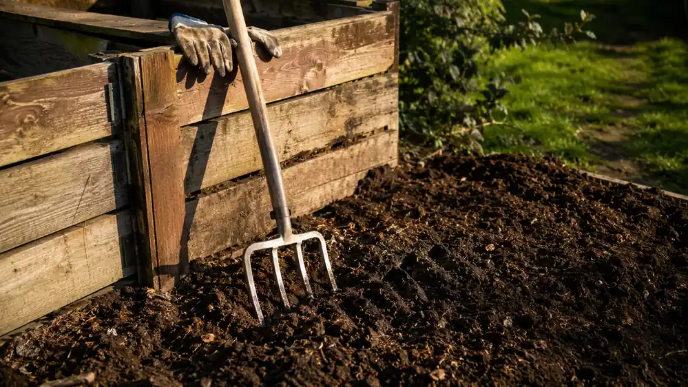 Spading fork leaning against a compost bin with turned compost and a garden fork's tine marks visible in the pile